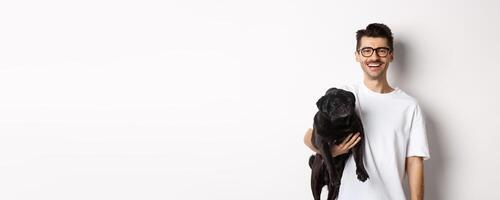 Handsome hipster guy holding his funny black pug dog, smiling at camera, standing over white background photo