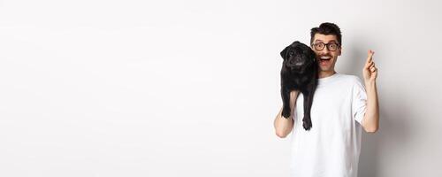 Hopeful smiling dog owner making a wish, holding cute black pug on shoulder and cross fingers for good luck, white background photo