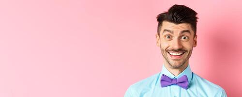Close-up of cheerful caucasian man smiling happy, looking at something interesting, standing in bow-tie over pink background photo