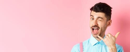 Cheeky and funny guy showing phone gesture, asking to call him and winking, telling dial his number, standing over pink background photo