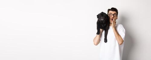 Image of shocked dog owner staring at camera and gasping impressed, cute black pug sitting on his shoulder and staring at you, white background photo