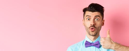 Close-up of funny guy with moustache, say wow and showing thumb up in approval, checking out something cool, recommending product, standing on pink background photo