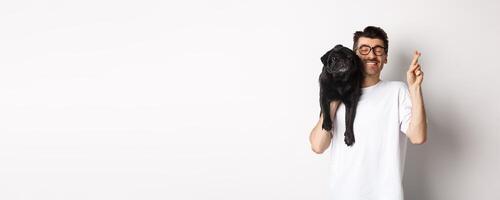 Hopeful smiling dog owner making a wish, holding cute black pug on shoulder and cross fingers for good luck, white background photo