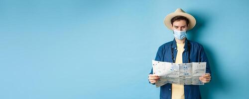 Covid-19, pandemic and travel concept. Young man in medical mask reading tourist map, searching for route to sightseeings, standing on blue background photo