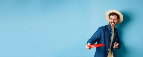 Happy man laughing and playing frisbee, throwing at friend and smiling, standing in summer hat on blue background photo