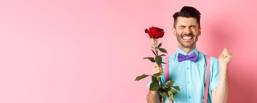 romance y san valentin día concepto. esperanzado hombre sensación emocionado antes de fecha, esperando para amante con rojo Rosa y Orando, en pie terminado rosado antecedentes foto