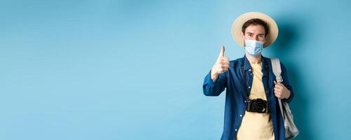 Covid-19 and summer holidays concept. Cheerful tourist in straw hat and medical mask praise and recommend good vacation hotel, showing thumb up, going on travel with backpack photo