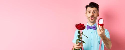 Valentines day. Funny man with moustache and bow-tie making proposal, showing engagement ring and propose with red rose, standing over pink background photo