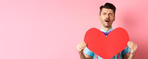 Valentines day concept. Sad and lonely man feeling heartbroken, being rejected, showing big red heart cutout and crying from break-up, standing on pink background photo