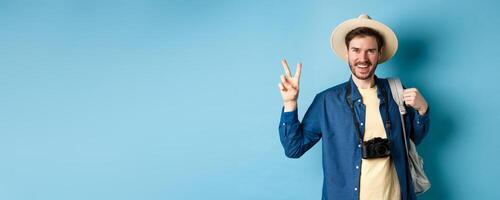 Happy handsome man taking photo on summer vacation, showing peace sign and smiling, wearing straw hat and holding tourist backpack, blue background