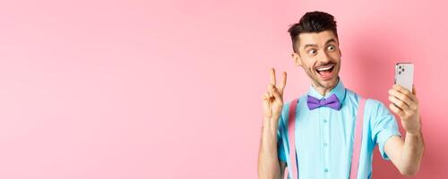 Technology concept. Funny man with moustache and bow-tie taking selfie on smartphone, showing peace sign and smiling at mobile camera, pink background photo