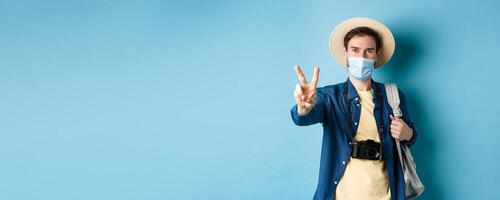 Covid-19, pandemic and travel concept. Positive guy going on vacation, wearing summer hat and medical mask, showing peace sign, standing with backpack and camera on blue background photo