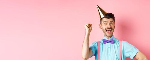 Holidays and celebration concept. Happy birthday boy with moustache and bow-tie celebrating event in party cone hat and festive clothes, raising hand up and shout yes photo
