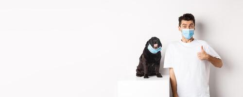 Covid-19, animals and quarantine concept. Image of funny young man and small dog in medical masks, owner showing thumb up in approval, praise something, white background photo
