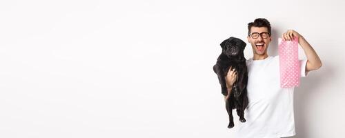 Image of happy man, pet owner, holding cute black pug and dog poop bag, standing over white background photo