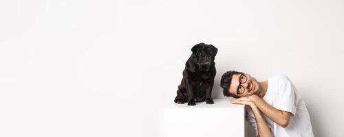 Handsome young man lay head near cute black pug, smiling and looking up at copy space, white background photo