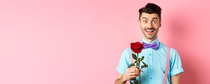 emocionado barbado hombre con Bigote y corbata de moño esperando para fecha con rojo rosa, teniendo romántico momento en san valentin día, en pie en rosado antecedentes foto