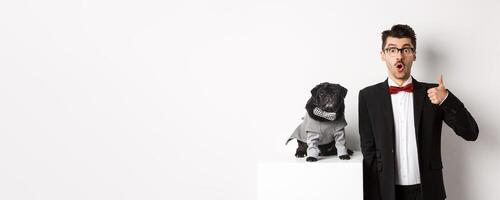 Animals, party and celebration concept. Handsome young man in suit and cute black pug in costume staring at camera, owner showing thumb up in approval and praise, white background photo