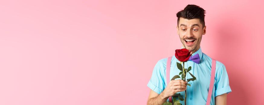 san valentin día y romance concepto. contento novio mirando a rojo Rosa mientras esperando para amante en corbata de moño, en pie en rosado romántico antecedentes foto