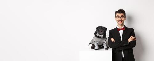 Animals, party and celebration concept. Happy dog owner in suit and puppy in costume looking excited at camera, having fun, standing over white background photo
