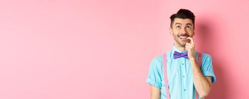 Image of smiling man making choice, looking dreamy and happy up, biting fingernail with tempted expression, wanting something, standing over pink background photo
