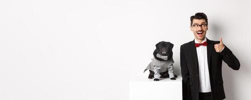 Animals, party and celebration concept. Handsome young man in suit and cute black pug in costume staring at camera, owner showing thumb up in approval and praise, white background photo