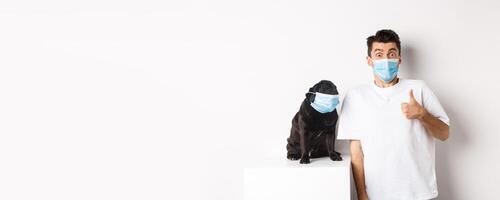 Covid-19, animals and quarantine concept. Image of funny young man and small dog in medical masks, owner showing thumb up in approval or like, white background photo