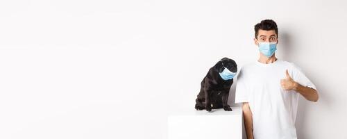 Covid-19, animals and quarantine concept. Image of funny young man and small dog in medical masks, owner showing thumb up in approval, praise something, white background photo