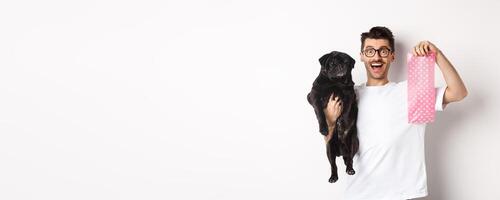 Image of hipster guy pet owner, holding cute black pug and dog poop bag, standing over white background photo