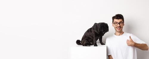 Image of happy and satisfied young man sitting near pug dog and showing thumbs-up, smiling and praising good product, white background photo