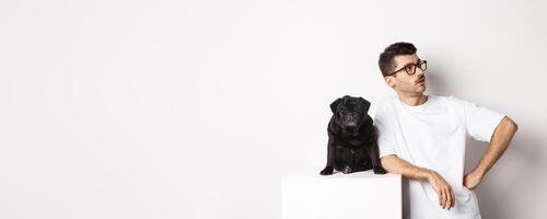 Handsome young man standing near cute black pug, looking right with arrogant expression, standing over white background photo