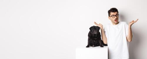 Portrait of confused hipster guy dog owner shrugging, standing near cute black pug pet, white background photo