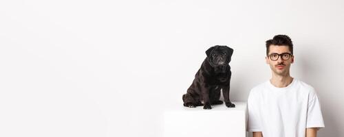 Image of handsome hipster in glasses sitting next to black cute pug dog, both staring at camera over white background photo