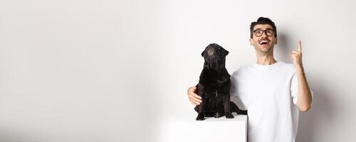 Image of happy young man and dog both looking up at promo, owner pointing finger at top, pug staring at loog, standing over white background photo