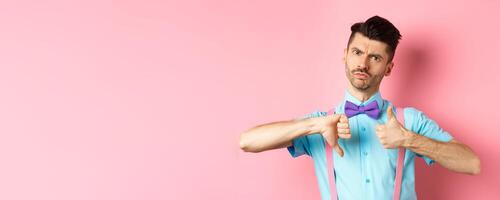 Skeptical young man in bow-tie grimacing, showing thumbs up and down, standing indecisive and judging something, standing over pink background photo