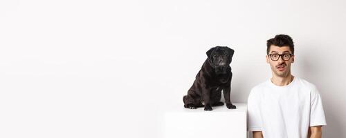 Image of hipster dog owner sitting near cute black pug, staring at camera confused and puzzled, white background photo