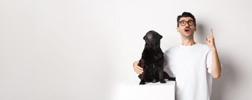 Image of happy young man and dog both looking up at promo, owner pointing finger at top, pug staring at loog, standing over white background photo