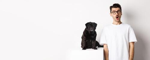 Image of dog owner and cute black pug looking at camera surprised and amazed, standing over white background photo
