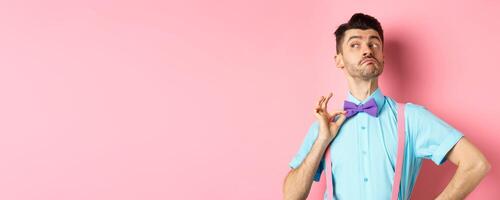 Image of funny man raising chin up and adjusting bow-tie, standing confident and sassy on pink background, looking left photo