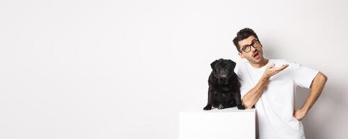 Image of hipster guy talking with his dog, looking skeptical and gesturing, having conversation with cute black pug, standing over white background photo
