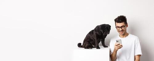 Smiling young man using smartphone and sitting near dog. Pug owner checking photos on mobile phone, white background