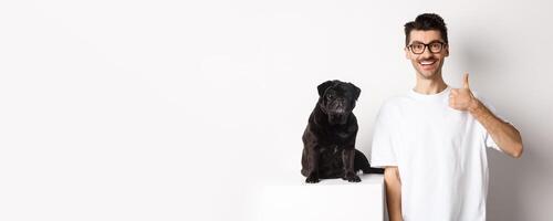 Image of dog owner and cute black pug looking at camera, man showing thumb-up in approval, recommending something, standing over white background photo
