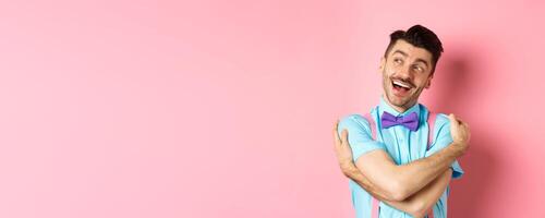 Happy and positive guy in bow-tie, hugging himself and looking left with cheerful smile, standing over pink background photo