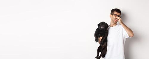 Dog owner squinting and shutting nose from disgusting reek, holding pug with bed smell, standing over white background photo