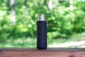 A black thermos with brewed tea or coffee stands on a wooden board with a copyspace. Thermos with hot drink photo