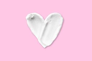Swatch Smear of cosmetic white cream in the shape of a heart isolated on a pink background. photo