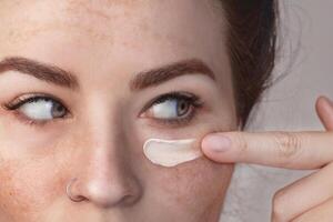 Caucasian woman's face with freckles or age spots, she smears a moisturizing, caring cream under the eye with her fingers, nice cream swatch, close up photo