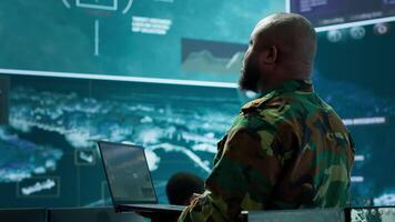 Military officer oversees surveillance and cybersecurity operations in a high tech army command post, ensuring the protection of classified information and data integrity. Big screen gear. Camera A. video