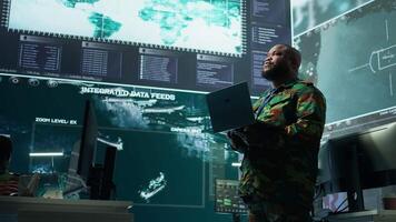 Military officer oversees surveillance and cybersecurity operations in a high tech army command post, ensuring the protection of classified information and data integrity. Big screen gear. Camera B. video