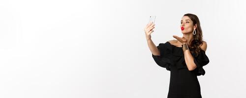 Stylish young woman in black dress, partying and taking selfie on mobile phone, sending air kiss at smartphone camera, standing over white background photo